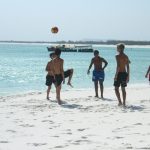 five boys playing football on a beach with a small boat in the background
