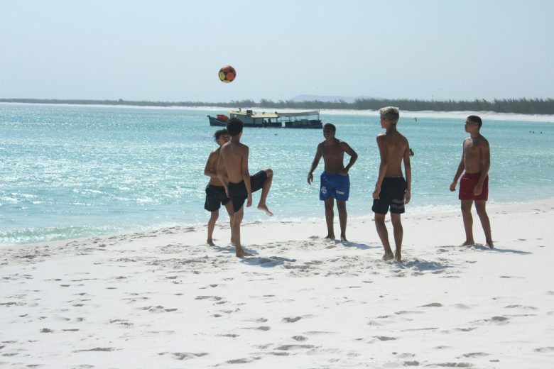 five boys playing football on a beach with a small boat in the background